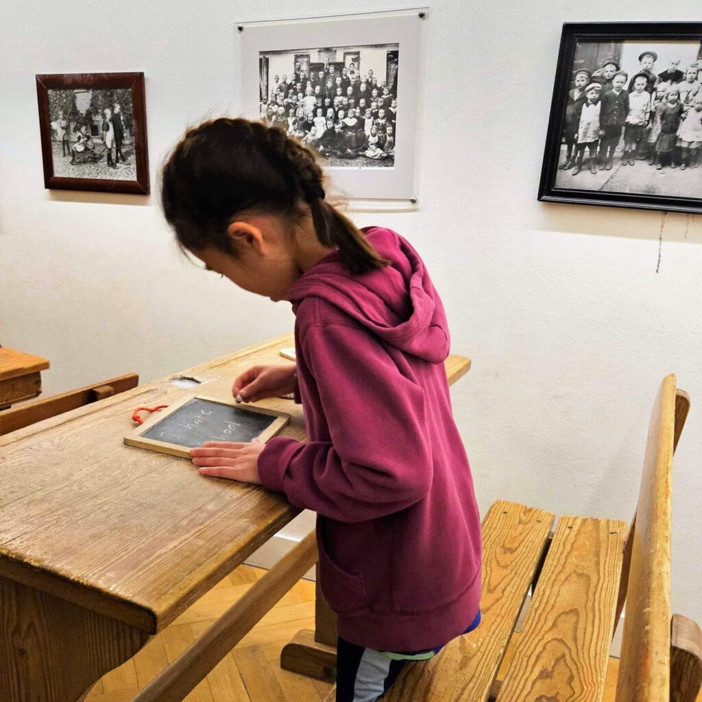 girl in purple sweatshirt leans over wooden desk; old photos are on the wall behind her