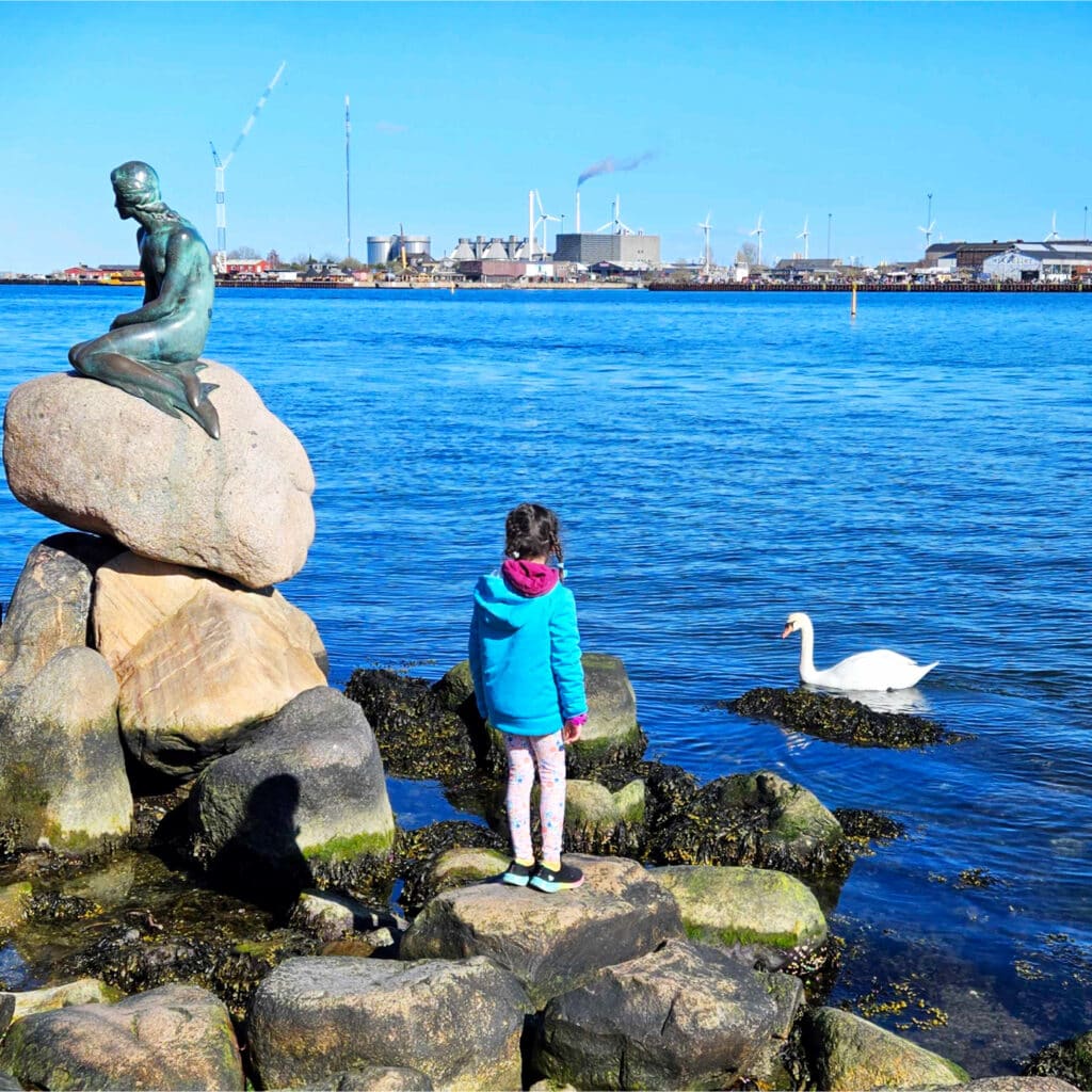 girl looks out at the river in Copenhagen; a mermaid (or merman) statue is to her left on top of a pile of large rocks