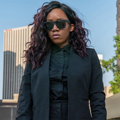 A woman wearing black sunglasses, a black blazer, and black blouse; a tall building is in the distance behind her