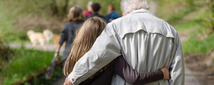 an old man and young girl walk together with their arms around each others' backs; he has white hair and a tan windbreaker jacket and she has long brown hair and a brown shirt; they are walking in a park