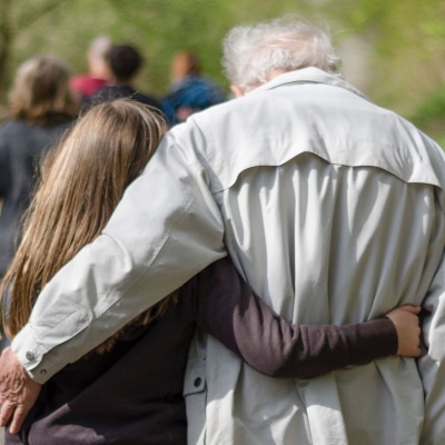 an old man and young girl walk together with their arms around each others' backs; he has white hair and a tan windbreaker jacket and she has long brown hair and a brown shirt; they are walking in a park