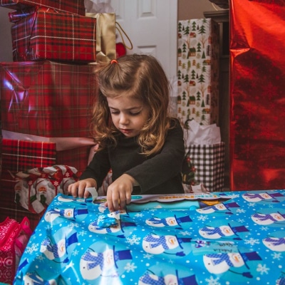 little girl opens a present wrapped with blue paper with snowmen on it; in the background are stacks of other presents wrapped in a variety of wrapping papers.