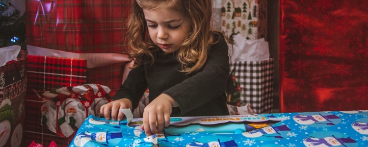 little girl opens a present wrapped with blue paper with snowmen on it; in the background are stacks of other presents wrapped in a variety of wrapping papers.