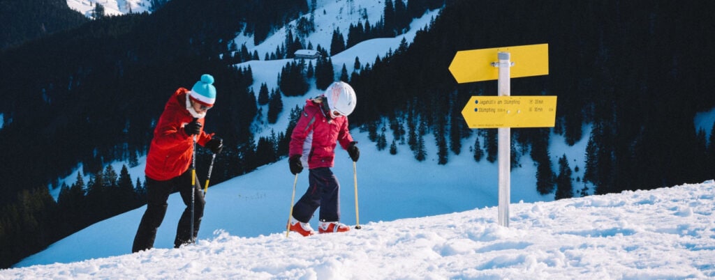 mother and child climb snowy hill with skiis and ski poles; there are mountains and trees in the background and 2 yellow wayfinder signs in front of them