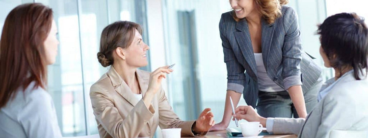 four women wearing blazers are chatting; three are seated and the fourth is leaning onto the table