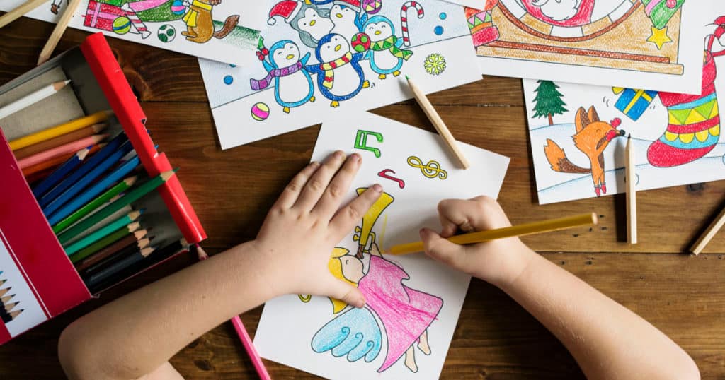 A child's hands coloring in a coloring book on a table, next to colored-in pages and a box of colored pencils