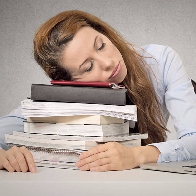 A woman sleeping on her table with her head on top of stacks of paperwork