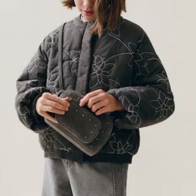young woman wears a quilted gray jacket with delicate white floral embroidery and light gray jeans