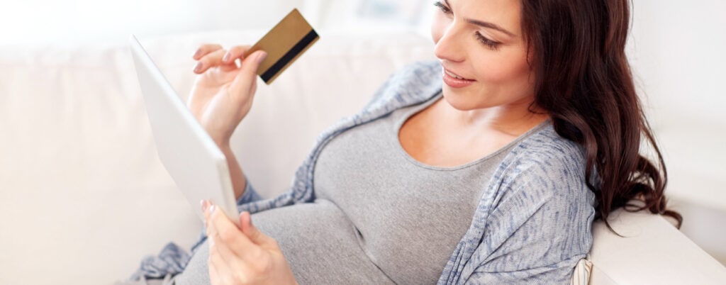 happy pregnant woman shopping on tablet with her credit card in her hand; she is apparently sitting on a beige couch in her apartment