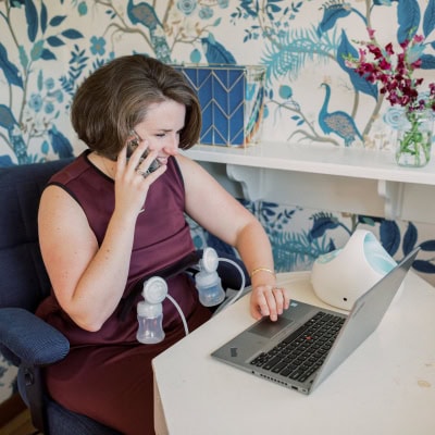working mother pumps breastmilk while talking on phone and typing on laptop; there is a blue wallpaper behind her with birds and leaves, as well as a blue chevron file box right behind her head and a vase filled with red flowers on a shelf