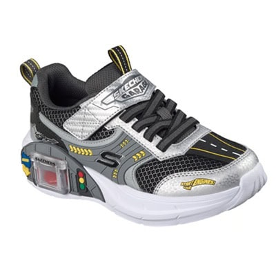 A kids' sneaker that is black, white, gray and yellow with a small "garage" in the side of the sole that contains a toy red car. Next to the garage is a traffic light.
