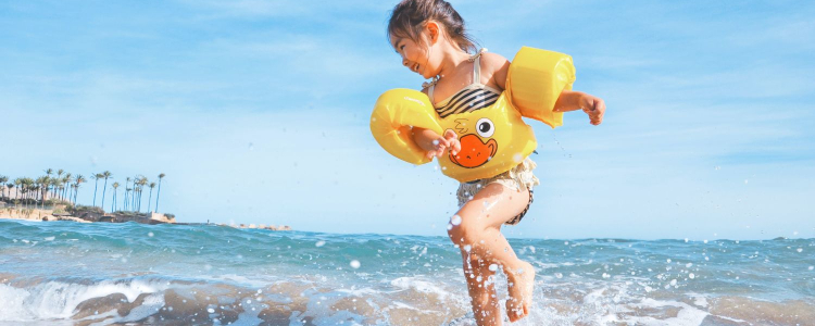 little girl in bathing suit and rubber duck floaty playing in ocean waves with palm trees in the distance