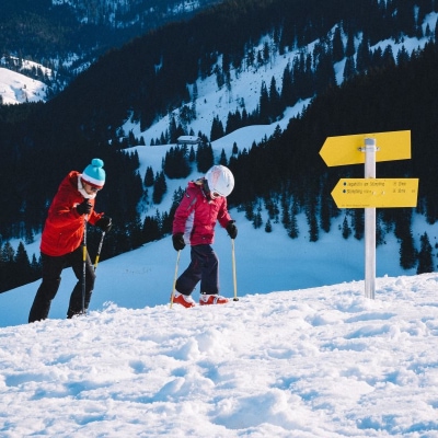mother and child climb snowy hill with skiis and ski poles; there are mountains and trees in the background and 2 yellow wayfinder signs in front of them