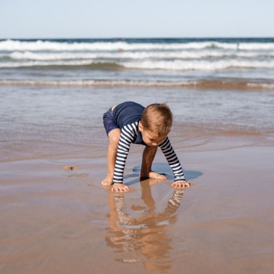 baby wears rash guard and plays in wet sand in front of ocean waves