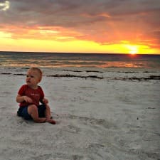 A baby sitting on the sand during sunset.
