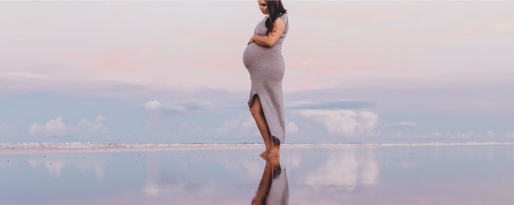pregnant woman in gray dress stands on reflective surface with a sunset and distant cityscape in the background