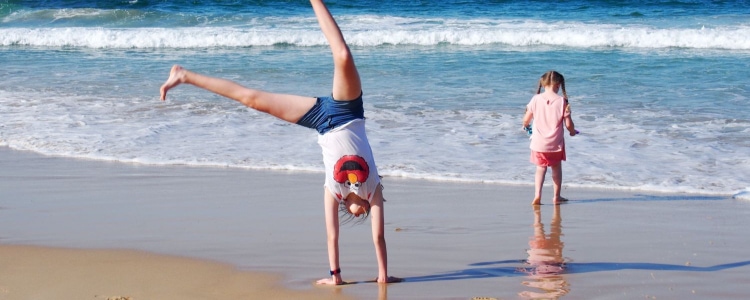 2 kids play on a beach; one is doing a cartwheel and another is close to wading into the ocean